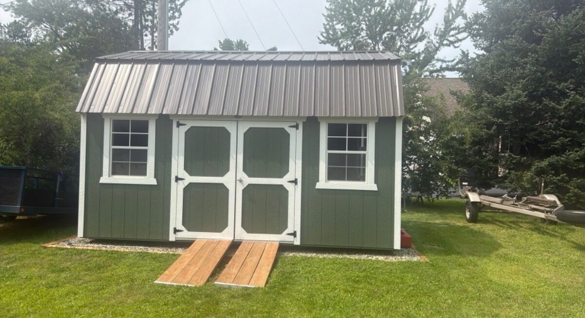 Green storage shed with white trim and a metal roof, set on a gravel base with wooden ramp rails leading to double doors